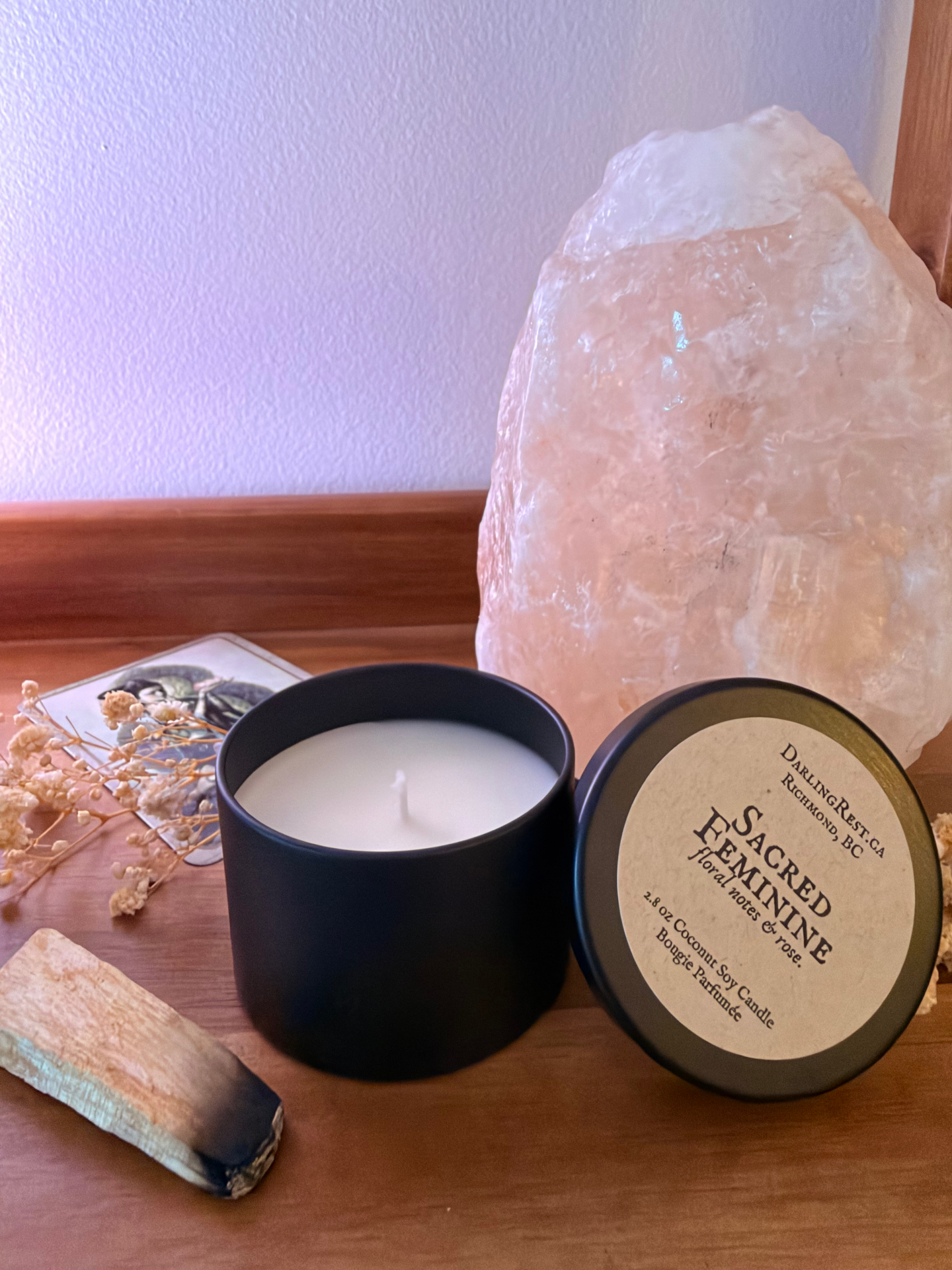 Sacred Feminine mini candle open beside its propped lid on a wooden table, with a pale pink crystal and dried flowers